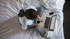 A woman sitting on her bed with her laptop and a cup of coffee.