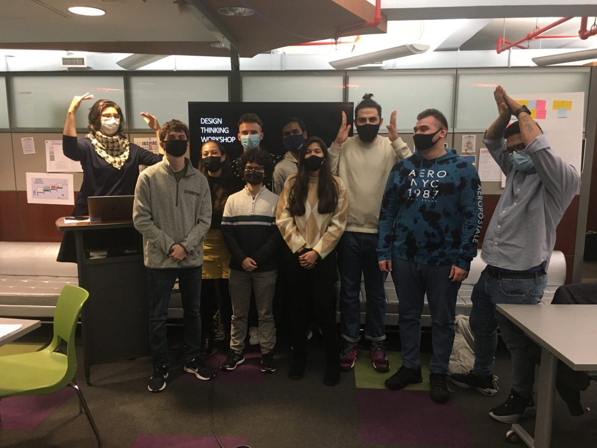 Seidenberg students pose in the Seidenberg Lounge NYC after completing the NYC Design Factory Workshop. Students are masked and are standing together for a group photo in front of a black powerpoint screen.